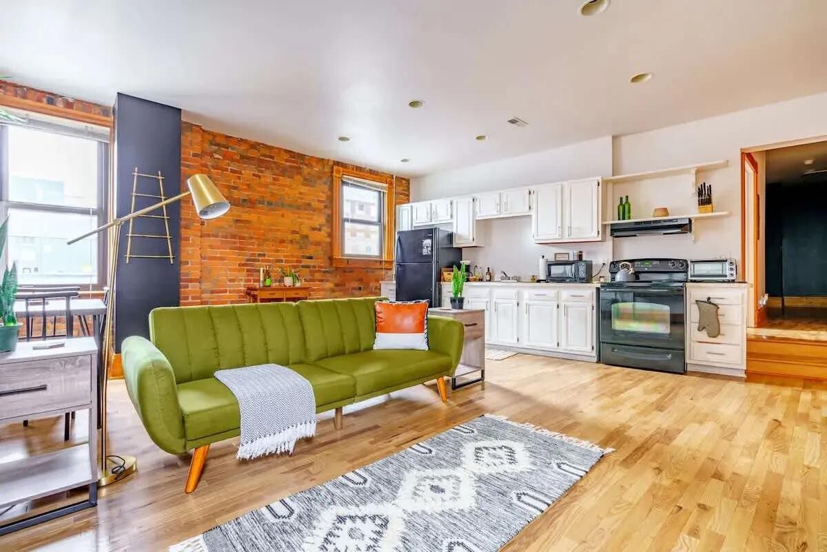 Luxury vacation rental living room featuring a green sofa, modern kitchen with white cabinetry, and exposed brick walls, ideal for visitors to Columbus seeking comfortable accommodations.